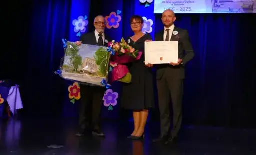 Obraz przedstawia trzy osoby na scenie SCK. W środku stoi kobieta z bukietem kwiatów. To dyrektorka SP nr 2 w Sztumie. Po jej dwóch stronach mężczyźni. Ten po prawej trzyma czek na 10 000 zł, a ten po lewej obraz.  To Burmistrz Sztumu i Przewodniczący Rad