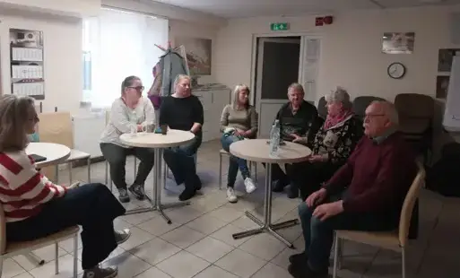 Zdjęcie przedstawia grupę osób prowadzących ożywioną dyskusję, skupioną wokół kilku okrągłych stolików. To grup wsparcia dla rodziców i opiekunów osób z zaburzeniami psychicznymi.